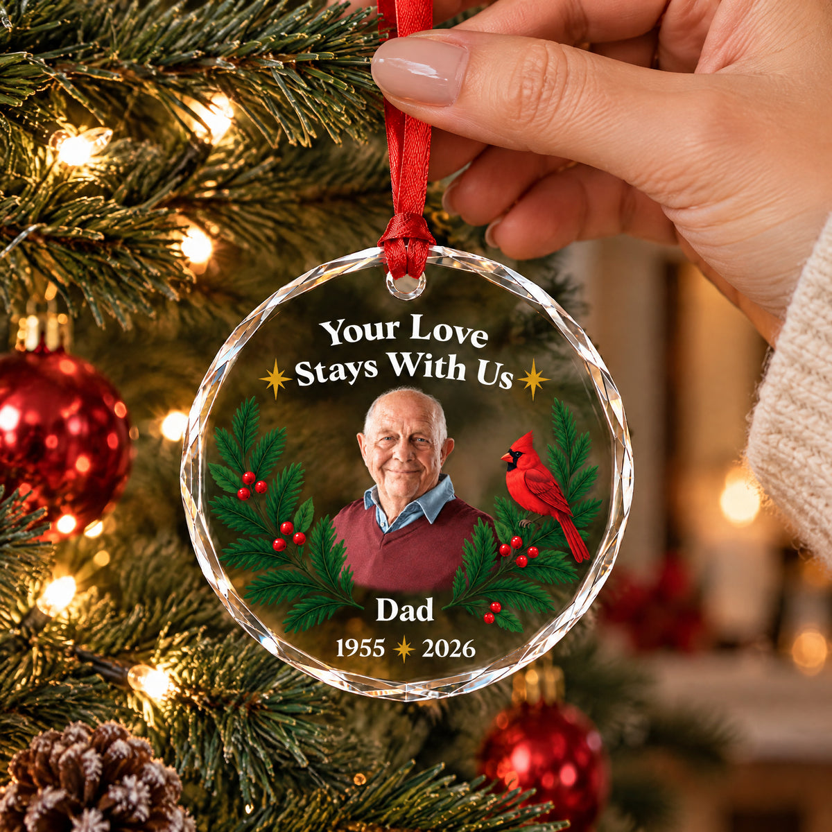 Personalized glass ornament custom photo held by a hand on a red ribbon in front of a decorated Christmas tree