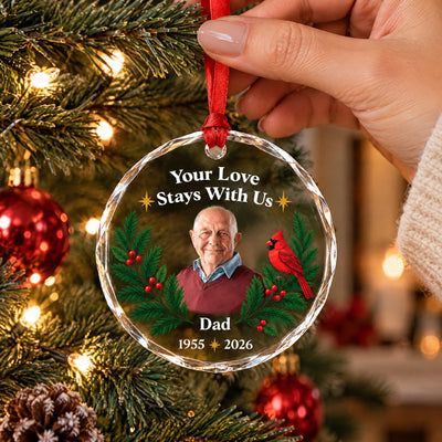 Personalized glass ornament custom photo held by a hand on a red ribbon in front of a decorated Christmas tree