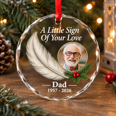 Personalized glass feather memorial ornament with photo and feather on a wooden table beside pinecones and pine branches