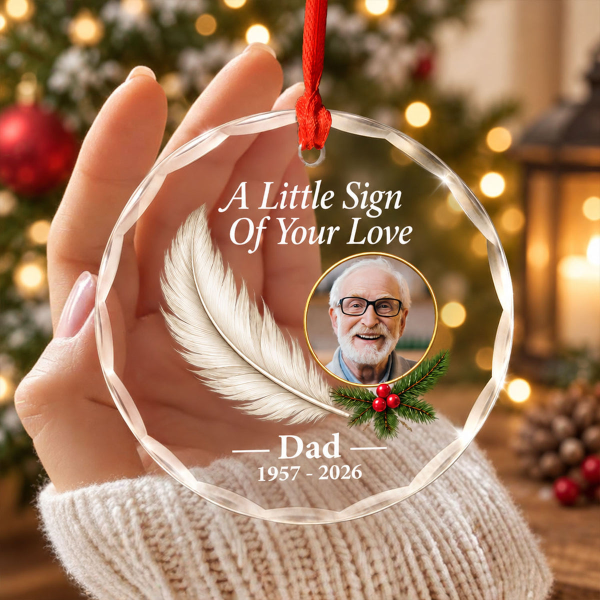 Personalized glass feather memorial ornament held in a hand near a blurred Christmas tree and lantern