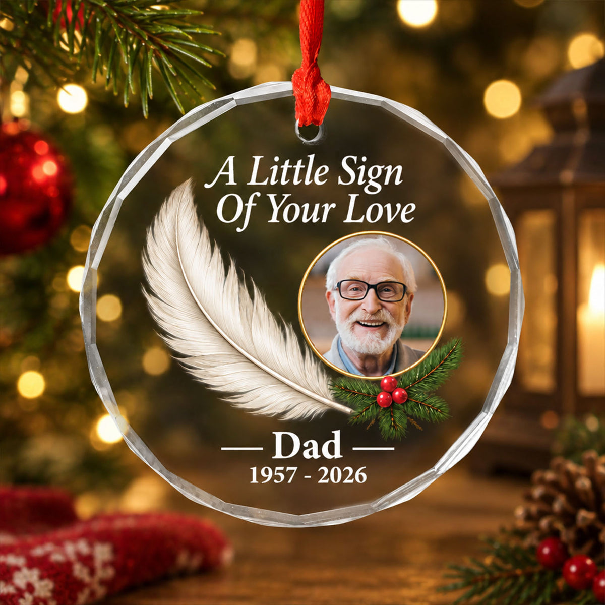 Personalized glass feather memorial ornament hanging by red ribbon in front of warm lights, a lantern and red baubles
