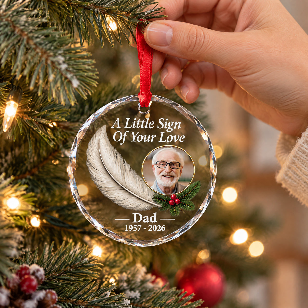 Personalized glass feather memorial ornament hung on a lit Christmas tree branch, held by a hand at the ribbon